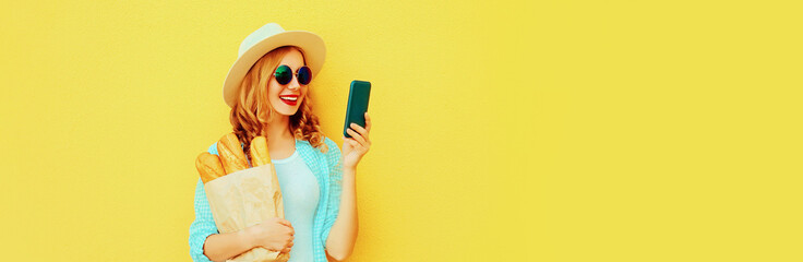Portrait of happy smiling woman with smartphone holding grocery shopping paper bag with long white bread baguette on yellow background, blank copy space for advertising text