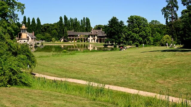 Versailles, Paris, France, June 2022. Nice pan footage of the Hameau de la Reine in the Petit Trianon park. People walk the paths in the middle of the meadows. Beautiful summer day.