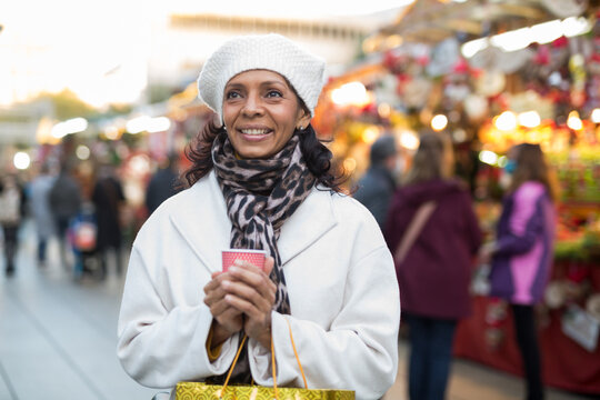 Cheerful Adult Latina Holding Paper Cup Of Hot Tea, Enjoying Walking At Street Fair Among Trade Stalls Offering Colorful Christmas Decorations