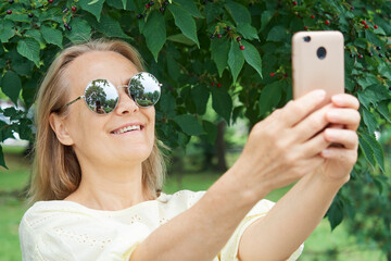 Close up of smiling mature lady in sunglasses taking selfie in park in summer