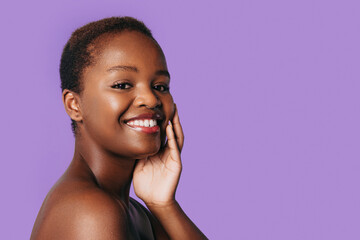 Portrait of smiling african american woman portrait looking at camera touching her healthy skin after spa procedure isolated over purple background with copy