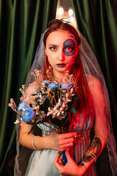 Beautiful girl in a blue dress with artistic makeup on her face holds a bouquet of flowers. Halloween mask. Red backlight on the side. Halloween concept, nightclub, masquerade