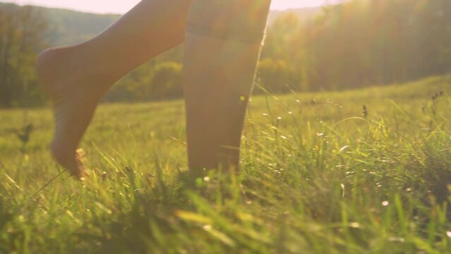 CLOSE UP, LOW ANGLE: Barefoot young woman walking on green grass in golden light. Carefree and relaxing moment in nature on a beautiful sunny morning. Female person pacing the meadow with bare feet.