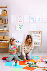 Portrait of attentive little girl sit with young woman teacher explaining how to play with wooden clock in classroom.