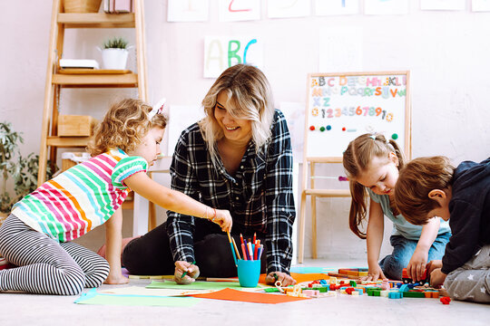 Portrait of young joyful woman teacher sit with children, looking at serious little girl taking pencil in classroom.