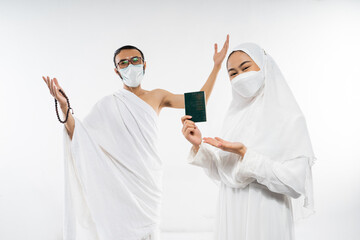 Happy couple in ihram clothes and mask holding a passport standing on isolated background