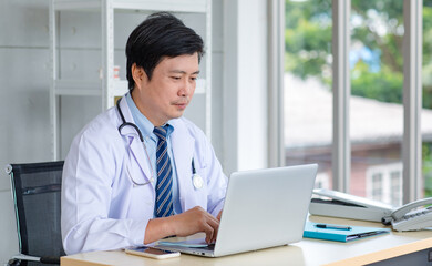 Asian middle aged male professional successful doctor practitioner in white lab coat with stethoscope sitting working at workstation desk typing laptop notebook computer in clinic hospital office - Powered by Adobe