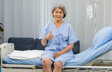 Portrait shot Asian old senior healthy gray hair female pensioner patient in blue hospital uniform sitting smiling holding hand showing thumb up on clinic bed in ward room with saline solution pole