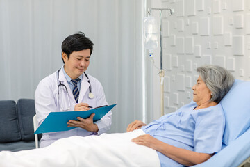 Asian male professional doctor practitioner in white lab coat and stethoscope writing notes when visiting monitoring old senior female pensioner grandma patient in hospital uniform laying down on bed