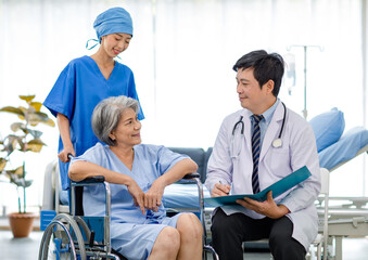 Asian male professional doctor practitioner in white lab coat and stethoscope writing notes when visiting monitoring examining old senior female pensioner patient sitting on wheelchair in hospital