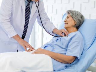 Asian male professional doctor practitioner in white lab coat using stethoscope listening examining heartbeat pulse of happy old senior female pensioner patient in hospital uniform lay down on bed