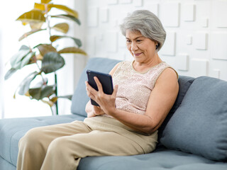 Asian cheerful old senior healthy gray hair female pensioner grandmother sitting smiling on cozy sofa in living room holding using touchscreen tablet computer surfing browsing application online