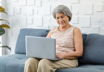 Asian cheerful old senior healthy gray hair female pensioner grandmother sitting smiling on cozy sofa in living room holding using laptop,notebook computer surfing browsing application online