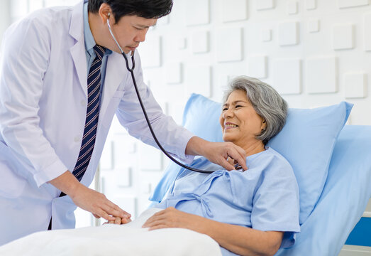 Asian male professional doctor practitioner in white lab coat using stethoscope listening examining heartbeat pulse of happy old senior female pensioner patient in hospital uniform lay down on bed