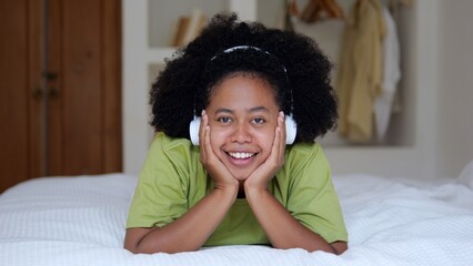 Fototapeta na wymiar A cute black woman with an afro hairstyle lies on the bed and smiles a snow-white smile. Afro Asian woman listens to music with wireless headphones. The life of a young multiracial woman.