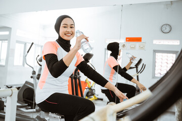 beautiful muslim woman drinking a bottle of water while exercising using static bike at the gym by...