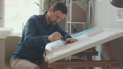 Side view medium slowmo of concentrated Caucasian male architect using compass while drawing building plan on desk in office - Powered by Adobe
