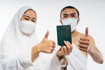 Man and woman wearing ihram clothes and mask holding a passport with thumbs up on isolated background