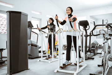 two beautiful woman with hijab at the gym exercising with friend on static elliptical cycle machine together - obrazy, fototapety, plakaty