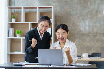 Two beautiful young Asian businesswomen raising their hands to express their joy at their satisfying accomplishment.