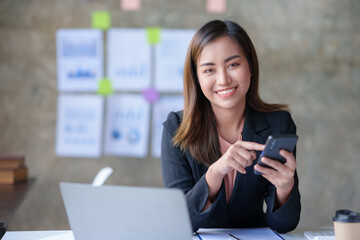 Beautiful Asian businesswoman using smartphone to view social media applications, respond to customer chats, make transactions through an application in the office.