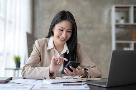 Beautiful Asian businesswoman using smartphone to view social media applications, respond to customer chats, make transactions through an application in the office.