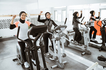 excited beautiful woman with hijab at the gym exercising with friend on static elliptical cycle...