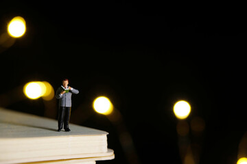 Miniature people toy figure photography. A boy reading a book at night with bokeh defocused light lamp.