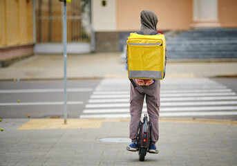Delivery boy riding electric unicycle, mono wheel with yellow backpack, delivery food service. Man from food delivery service with backpack on his back rides unicycle and delivers food. © Tricky Shark