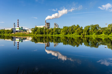 pipes of woodworking enterprise plant sawmill with beautiful reflection in blue water of river. Air pollution concept. Industrial landscape environmental pollution waste of thermal power plant