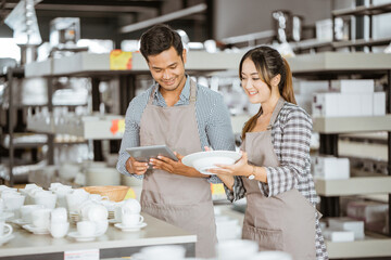 Man in apron using digital tablet and woman in apron holding plate in houseware store