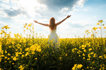Woman in a long dress stands in a field with yellow flowers and raising her hands to the sun.