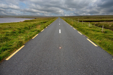 Small country road between a lake and green fields. Low cloudy sky. Travel and transportation industry.