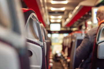Row of seats in a tourist bus and male passenger out of focus. Selective focus. Travel and commute concept. The interior has bald red color scheme, plastic seats and TV screen.