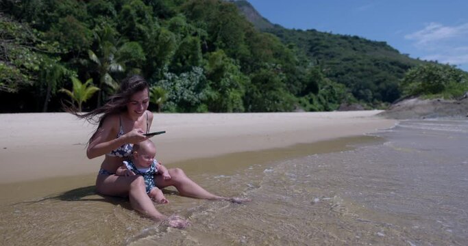 Young Mom takes cell phone photo of baby daughter on tropical beach - social media vacationing