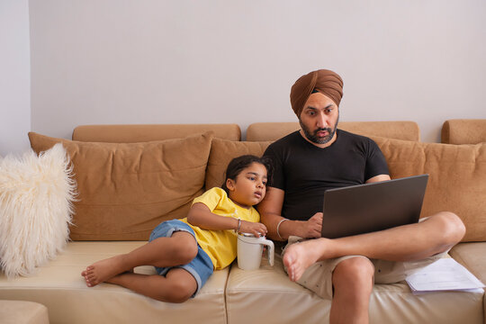 Sikh man working on laptop while daughter lying beside him on sofa