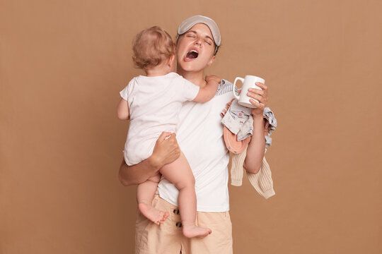 Tired Mother Yawning And Feels Tired, Holding Baby Stuff On Her Shoulder, Standing Indoor Isolated Over Brown Wall, Holding Cup Of Coffee And Keeps Eyes Closed.