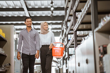 Asian muslim young couple carrying baskets walking between shelves in houseware store