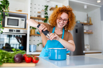 Cute young woman cooking and adding spice to meal, laughing and spending time in the kitchen