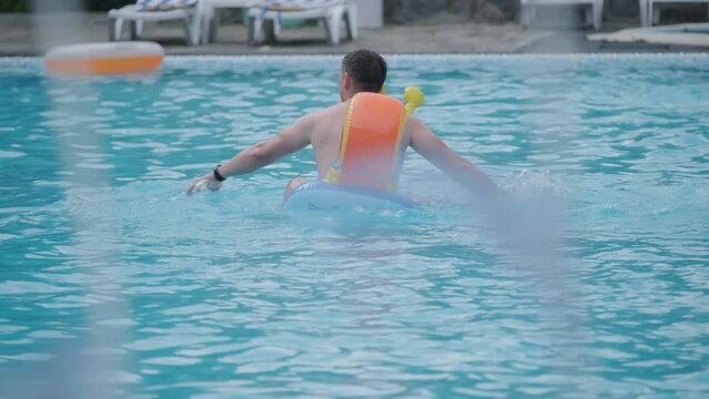 A funny man in an inflatable circle in the pool imitates swimming. View from behind. 4k video