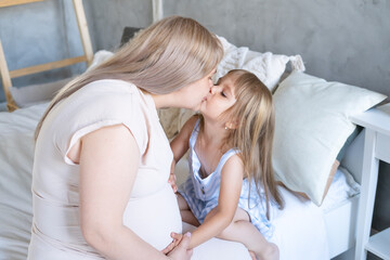 Pregnant woman sitting on bed with little daughter stroking tummy and kissing mother