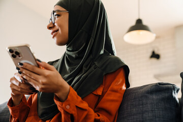 Young beautiful smiling woman in hijab with phone on sofa