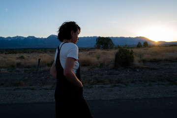 Pensive looking non-binary person wearing overalls on road trip walks in rural landscape with mountains at sunset