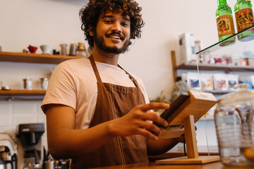 Young indian smiling handsome curly barista working with tablet