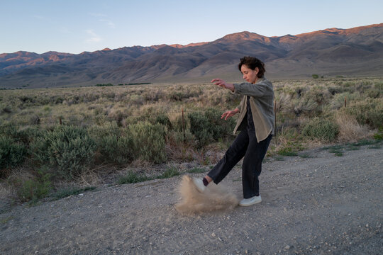Bored traveler kicks up dust on side of gravel road in desolate rural landscape