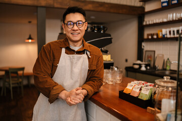 Adult asian man wearing apron smiling while working in cafe indoors