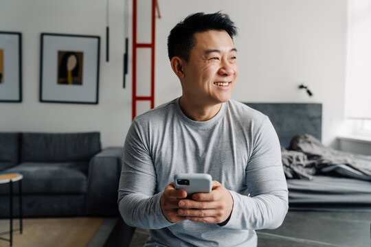 Brunette adult asian man smiling and using cellphone at home - Powered by Adobe