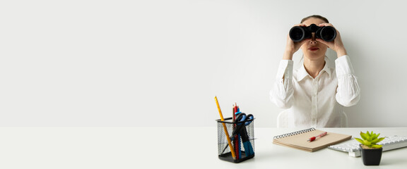 Young woman sitting in the office at the workplace looking through binoculars. Banner