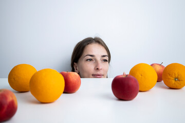 Fototapeta na wymiar Young woman looks at fruits lying on a white table against a white wall