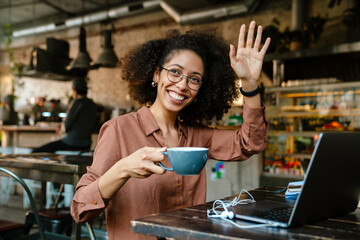 Young african american woman waving and drinking coffee in cafe